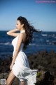 A woman in a white dress standing on a rock by the ocean.