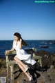 A woman in a white dress sitting on a bench by the ocean.