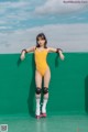 A woman in a yellow bodysuit on roller skates leaning against a green wall.