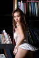 A woman in a white dress standing in front of a bookshelf.